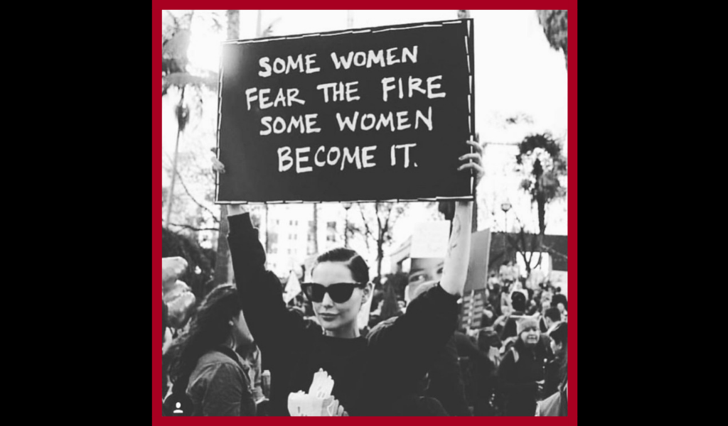 Black and white photo of a woman holding a protest sign that reads "Some women fear the fire. Some women become it."